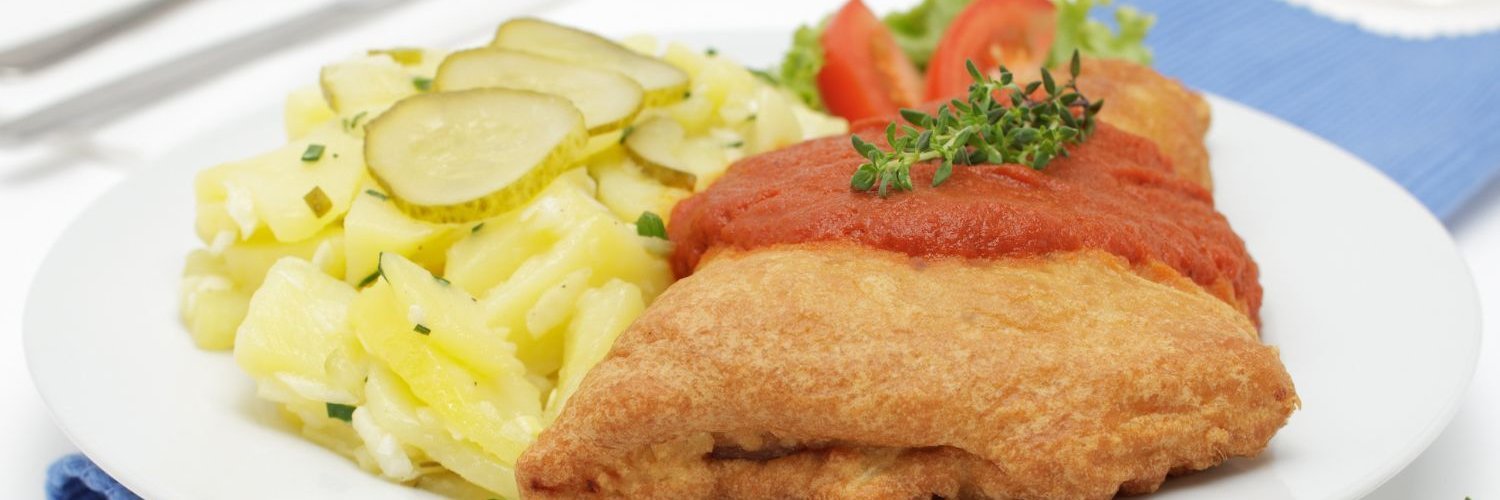 Auf einem weißen Teller liegt ein Stück paniertes Fischfilet mit Tomatensauce, daneben Kartoffelsalat mit Gurkenscheiben.