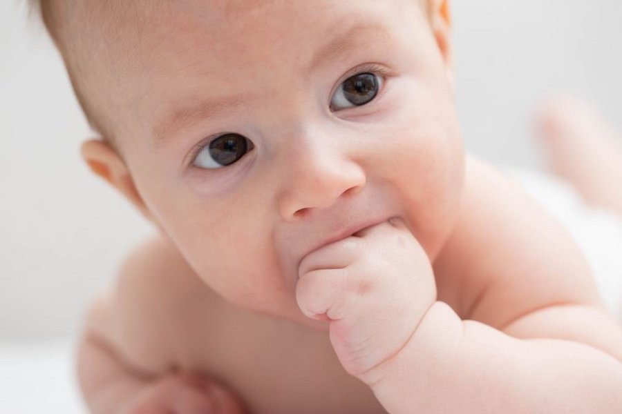 Ein auf dem Bauch liegendes und sich aufstützendes Baby hat seine Faust im Mund.