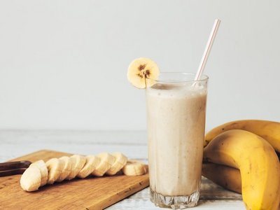 Eine Bananenmilch mit Strohhalm sowie ganzen und geschnittenen Bananen auf einem Schneidebrett..