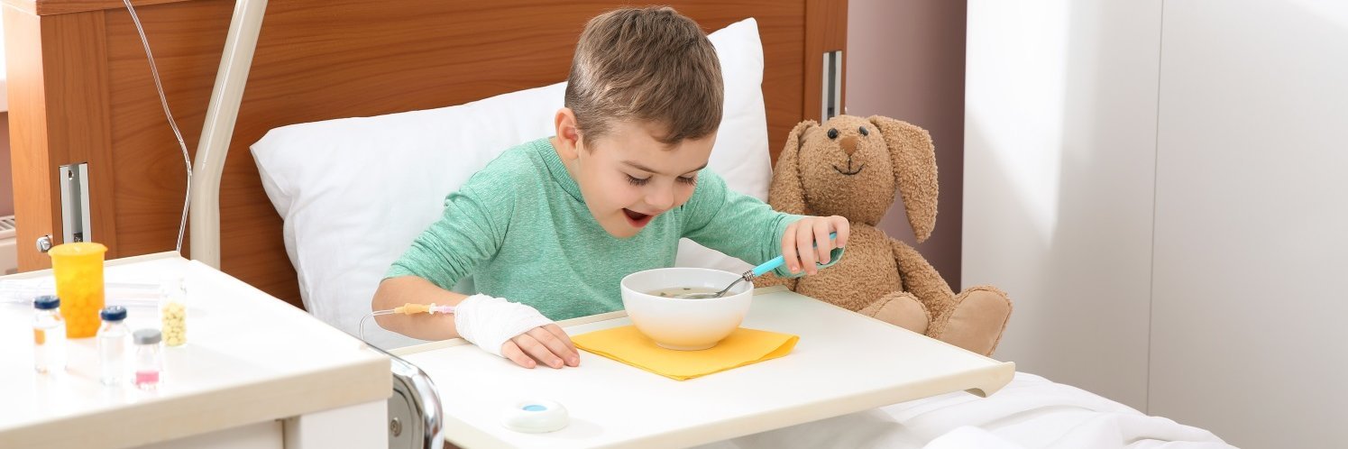 Ein kleiner Junge in grünem Oberteil mit Infusionszugang in der Hand sitzt in einem Klinikbett und isst Suppe aus einer Schüssel von dem Tabletttisch vor ihm. Neben ihm sitzt ein Plüschhund.