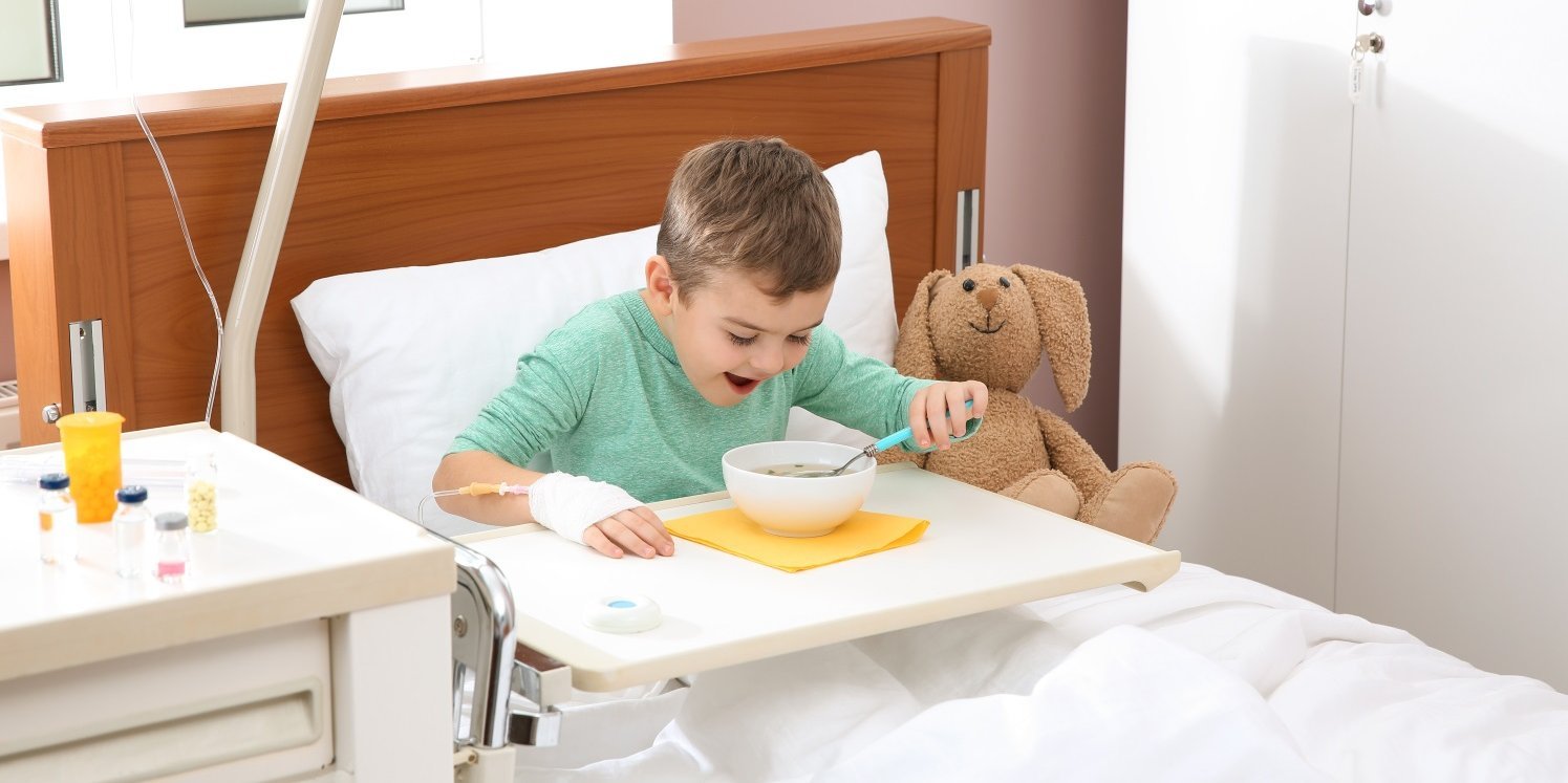 Ein kleiner Junge in grünem Oberteil mit Infusionszugang in der Hand sitzt in einem Klinikbett und isst Suppe aus einer Schüssel von dem Tabletttisch vor ihm. Neben ihm sitzt ein Plüschhund.. Artikel "Gesundes und nachhaltiges Essen für die Kinderklinik" öffnen.
