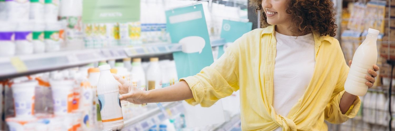 Eine junge dunkelhaarige Frau mit Einkaufswagen hält im Supermarkt eine Flasche Milch in der einen Hand und nimmt mit der anderen ein Milchprodukt aus dem Regal.