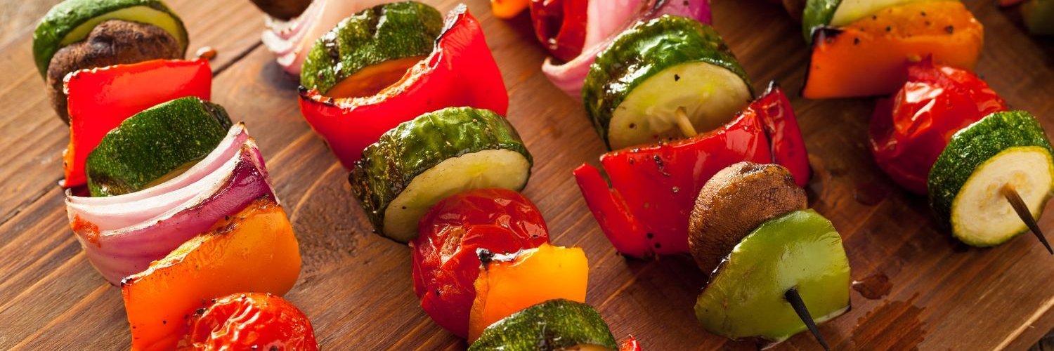 Auf einem Holzbrett liegen Spieße mit verschiedenen Gemüsestücken, wie Paprika, Zucchini und Zwiebeln.