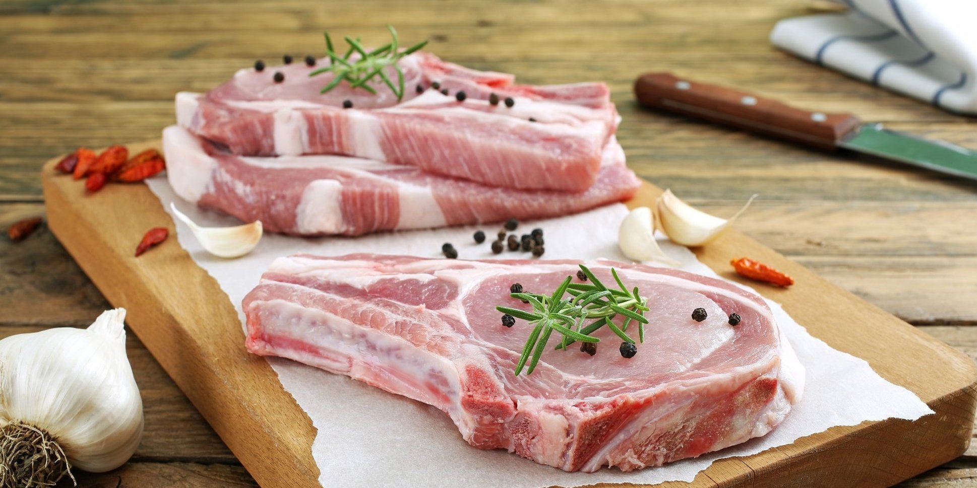 Rohes Schweinekotelett liegt mit Knoblauch, Pfefferkörnern, Chilischoten und Rosmarin auf einem Holzbrett.. Artikel "Schweinefleisch" öffnen.