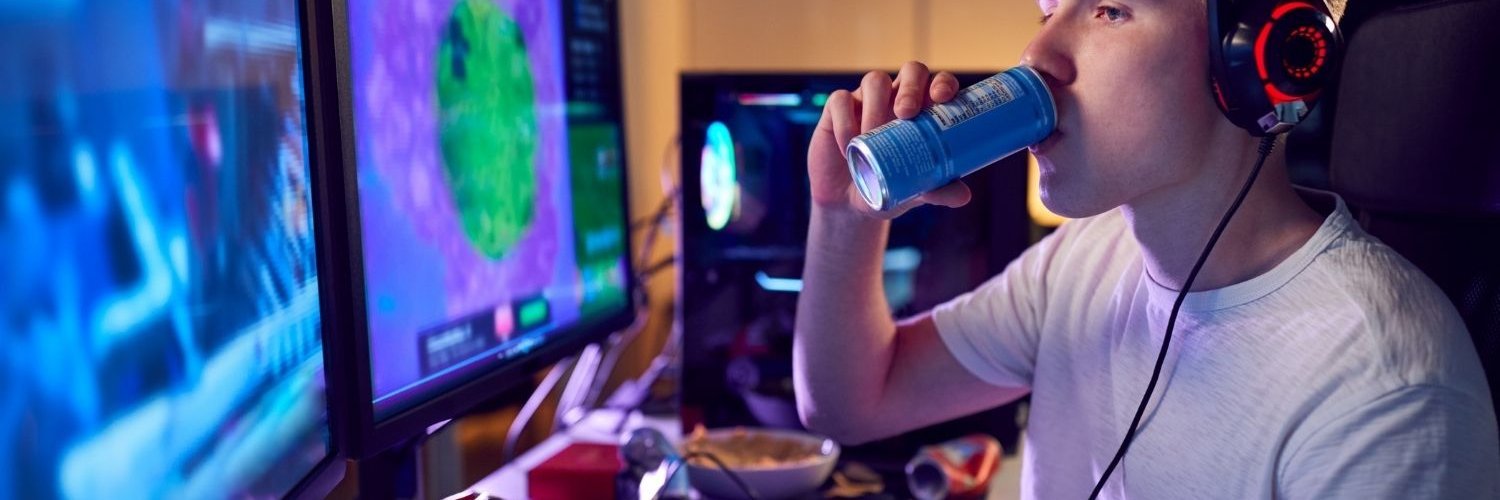 in blonder Jugendlicher im weißen T-Shirt mit Kopfhörern sitzt vor mehreren Bildschirmen und trinkt aus einer Dose.