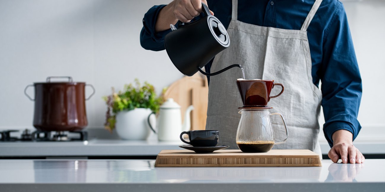 Ein Mann gießt Wasser aus einer Kanne in einen Handfilter für eine Tasse Kaffee.. Artikel "Kaffee: Vom Einkauf in die Küche" öffnen.