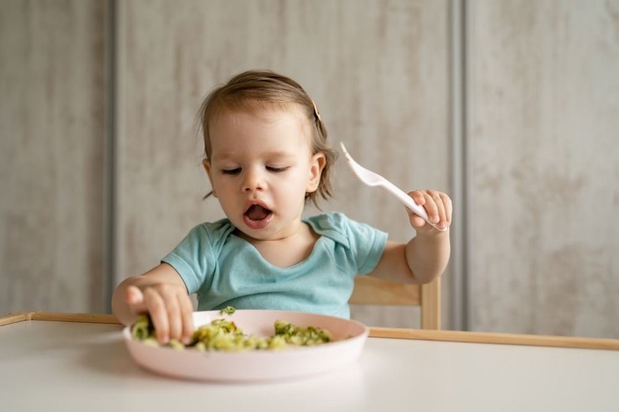 Eine Einjährige sitzt vor ihrem Teller und greift mit einer Hand nach gekochtem grünem Gemüse.