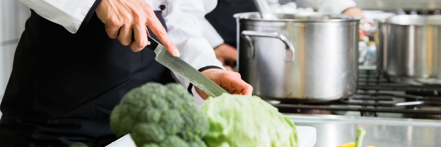 In einer Großküche arbeiten zwei Küchenkräfte an einem Stahltisch, eine schneidet Gemüse, die andere hält einen Schneebesen über einen Topf. 
