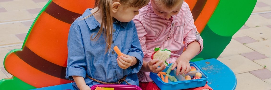 Ein Junge und ein Mädchen sitzen nebeneinander auf dem Schulhof und vergleichen ihr Pausenfrühstück..
