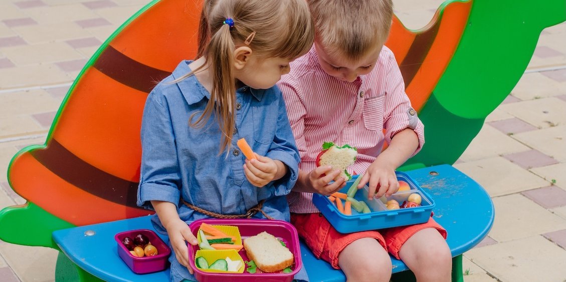 Ein Junge und ein Mädchen sitzen nebeneinander auf dem Schulhof und vergleichen ihr Pausenfrühstück..