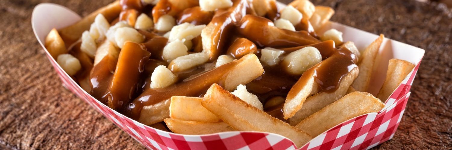 Auf einem groben Holztisch steht eine rot-weiß karierte Pappschachtel mit Pommes frites, die mit Bratensoße und Käse bedeckt sind.