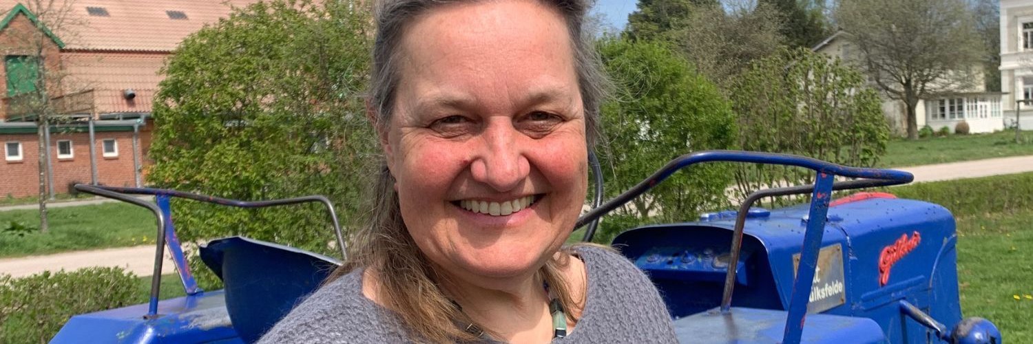 Eine dunkelhaarige Frau mittleren Alters steht im Freien vor einem Traktor und schaut in die Kamera, im Hintergrund sind landwirtschaftliche Gebäude.