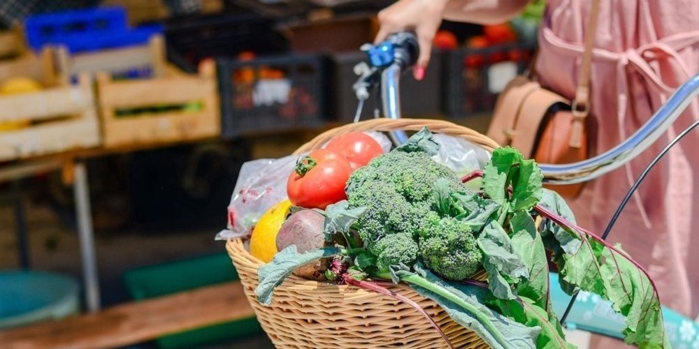 Eine Frau schiebt ihr Fahrrad auf dem Markt. Am Lenker hängt ein Korb gefüllt mit Brokkoli, Tomaten und Paprika. . Artikel "Gesund und nachhaltig essen mit kleinem Budget" öffnen.