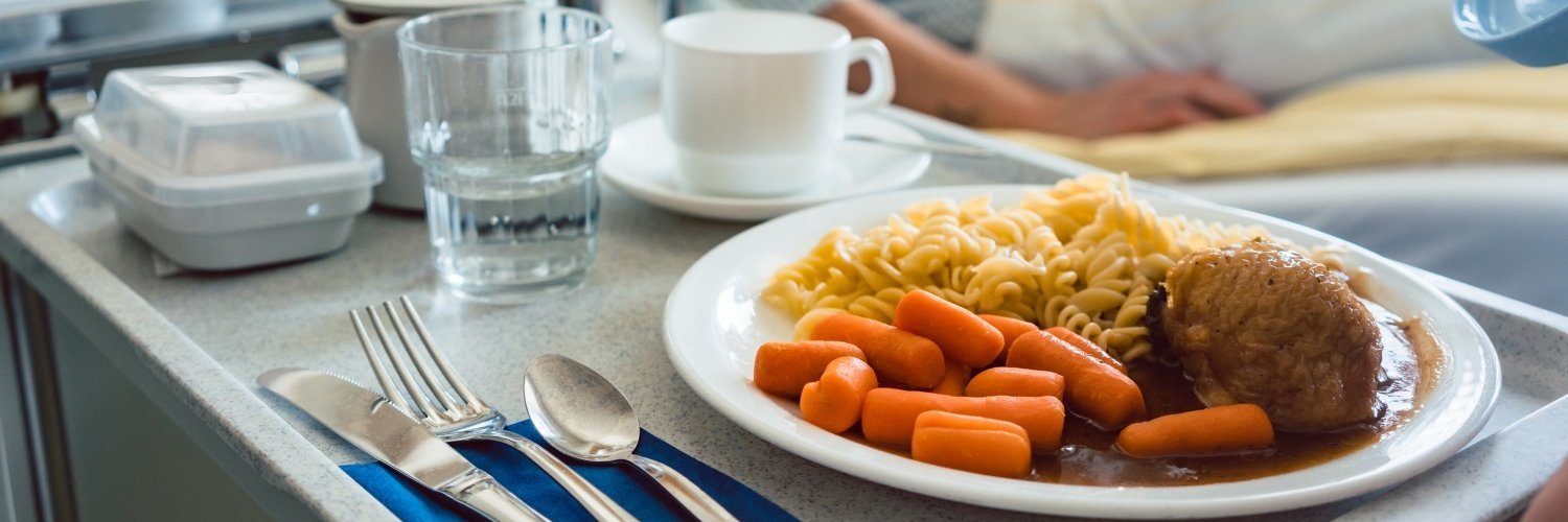 Auf einem Krankenhaustablett steht ein weißer Teller mit Möhren, Fleisch und Nudeln, ein Wasserglas, eine Tasse und eine kleine Teekanne, außerdem eine Dose. Daneben liegt ein Besteck auf einer Serviette. Im Hintergrund liegt eine Person im Bett.