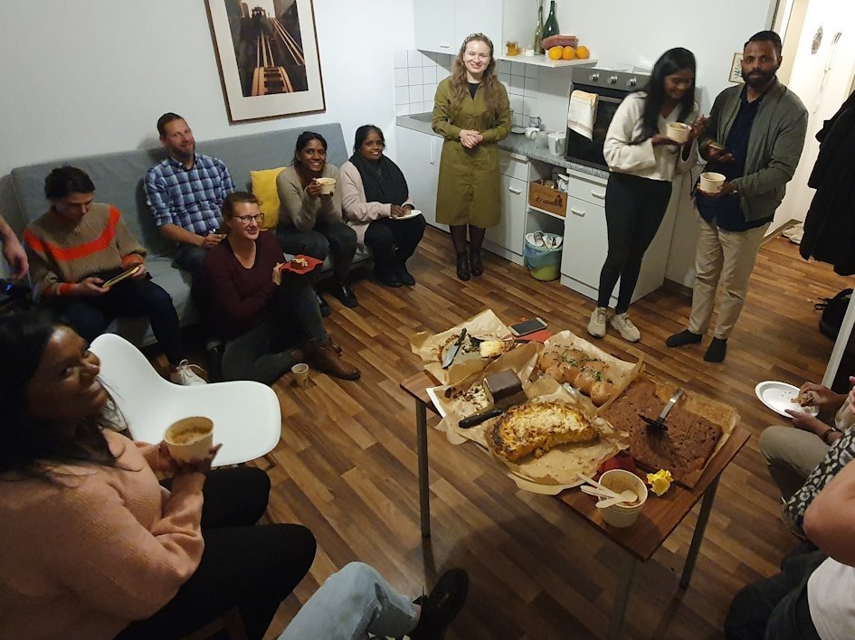 In einem Zimmer sitzen und stehen Menschen verschiedener Hautfarbe. In der Mitte steht ein Tisch mit Kuchen.
