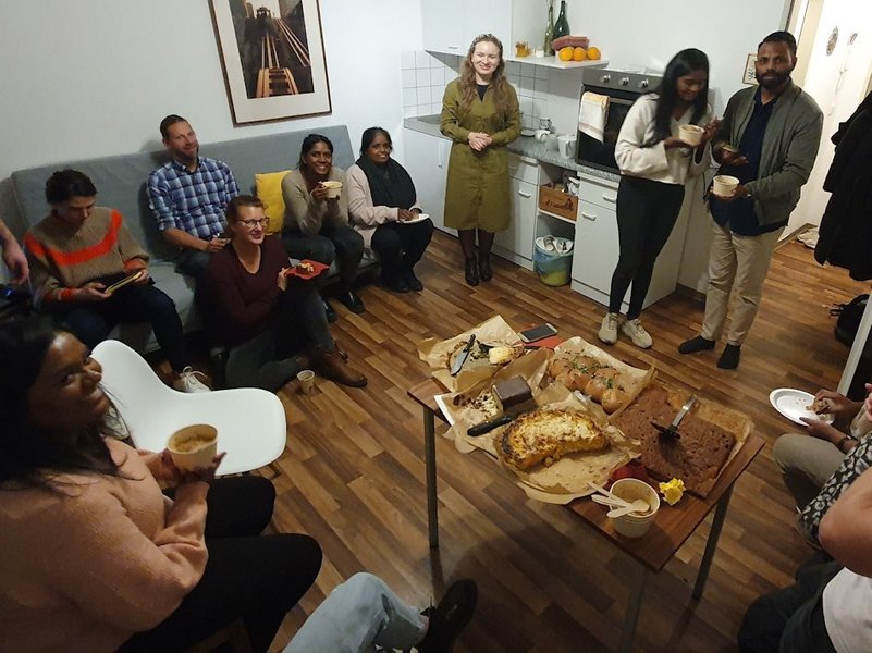 In einem Zimmer sitzen und stehen Menschen verschiedener Hautfarbe. In der Mitte steht ein Tisch mit Kuchen.