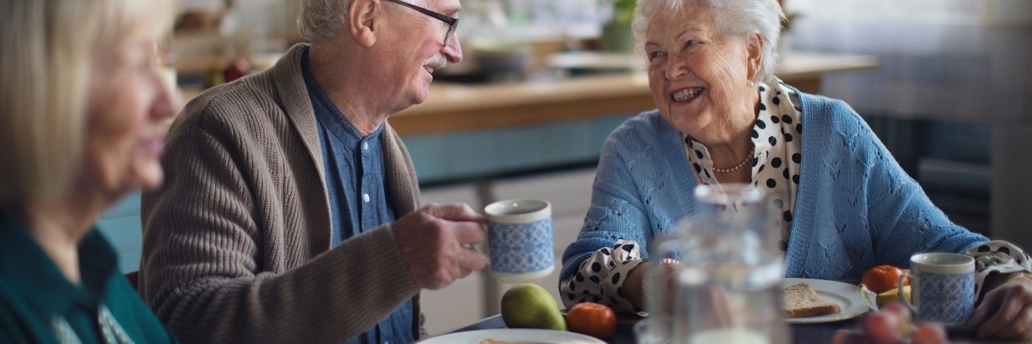 An einem Frühstückstisch sitzen Senioren und Seniorinnen zusammen, eine Frau und ein Mann lächeln einander zu.