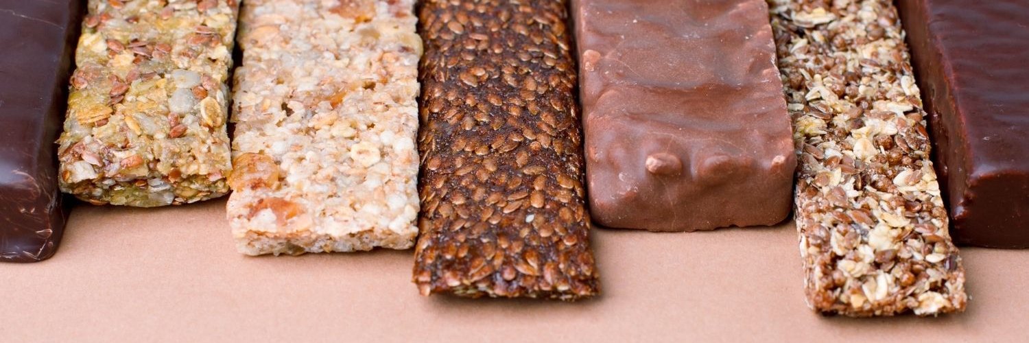 Aufgereiht nebeneinander liegen verschiedene Protein-, Schokoladen- und Müsliriegel am oberen Bildrand.