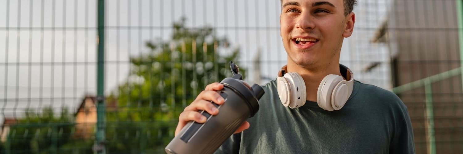 Ein Jugendlicher im T-Shirt und mit Kopfhörern um den Hals, steht vor einem Sportplatz und trinkt aus einem To-Go-Becher. 