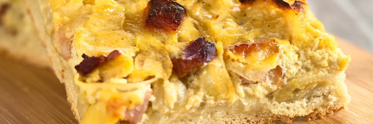 Auf einem Holzbrett liegt ein quadratisches Stück klassischer Zwiebelkuchen mit Speckstückchen.