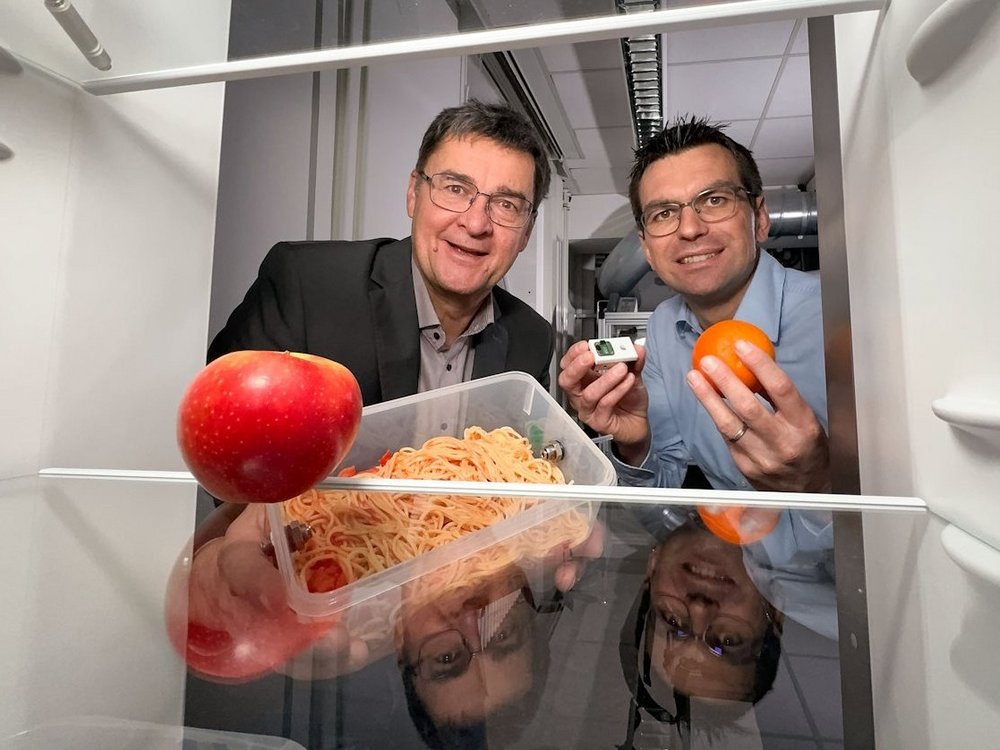 Der Blick aus einem offenen Kuehlschrank auf zwei davor sitzende Maenner, von denen einer ein Gefaess mit Spaghetti haelt und einer einen Computerchip und eine Orange.