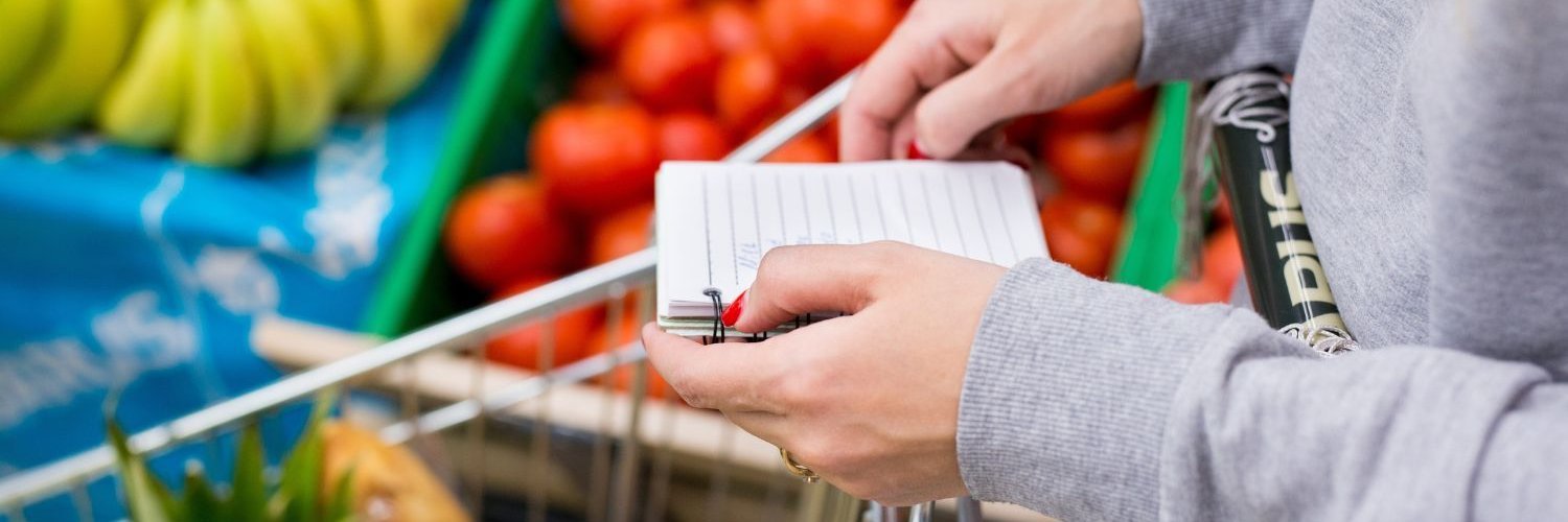 Eine Frau im grauen Sweatshirt schiebt einen Einkaufswagen durch die Gemüseabteilung und hält einen Block mit Notizen in der Hand.