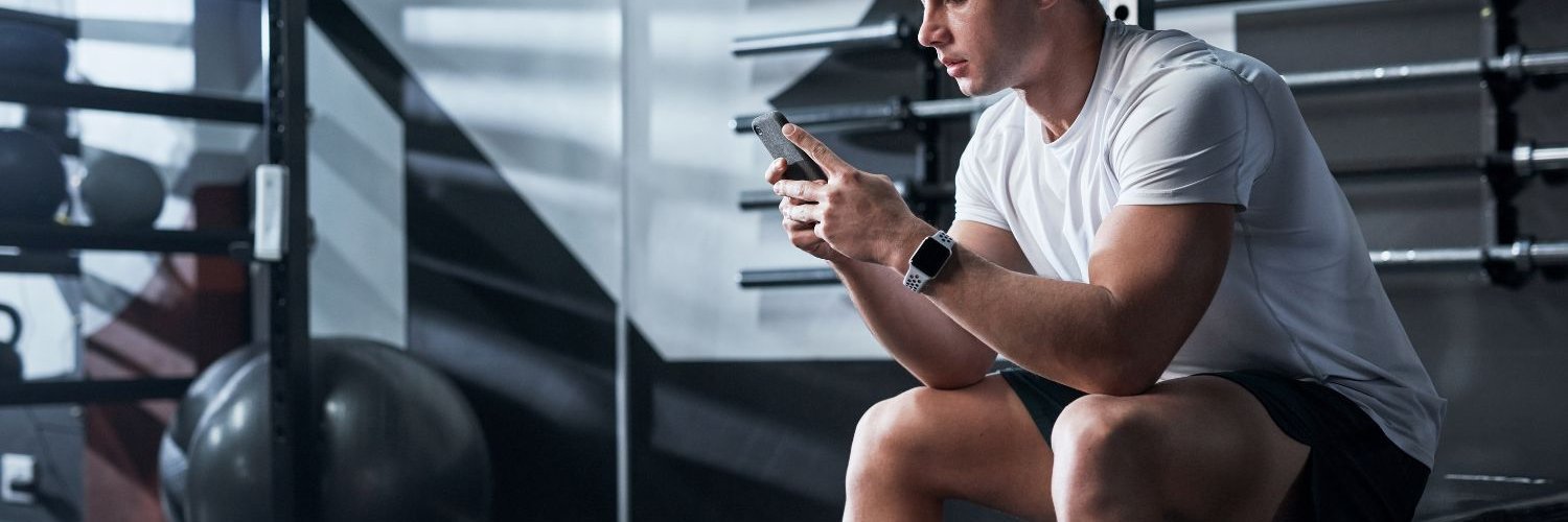 Ein junger Mann sitzt auf einem Trainingsgerät im Fitnessstudio und schaut besorgt auf sein Handy. 