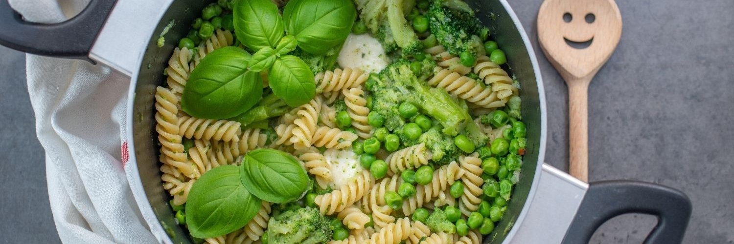Auf einem grauen Tisch steht ein Topf mit gekochten Spiralnudeln, grünen Erbsen, Brokkoli, Mozzarella und frischem Basilikum garniert.