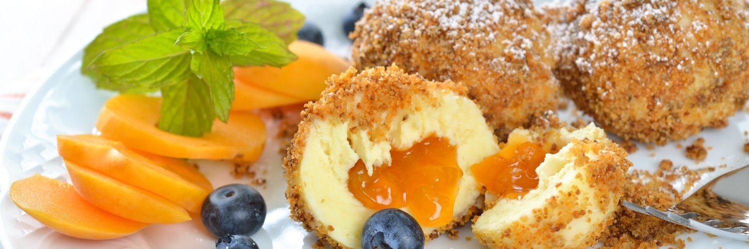 Auf einem weißen Teller liegen Marillenknödel mit Puderzucker. Einer ist halbiert und daneben liegen Aprikosenstücke und Blaubeeren.