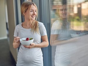 Eine schwangere Frau lehnt an eine Glastür und hält eine Schüssel mit Obstsalat in den Händen.