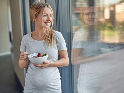 Eine schwangere Frau lehnt an eine Glastür und hält eine Schüssel mit Obstsalat in den Händen.