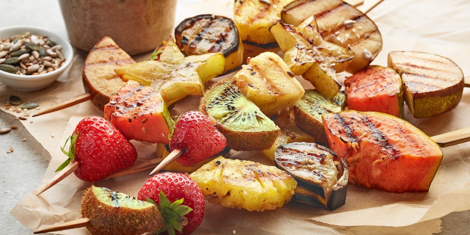 : Gegrillte Holzspieße mit Erdbeeren und Stücken von Kiwi, Birne, Melone, Ananas und Sternfrucht liegen auf Backpapier auf einem Tisch, daneben eine Schale mit Kürbis- und Sonnenblumenkernen.. Artikel "Süßes vom Grill" öffnen.