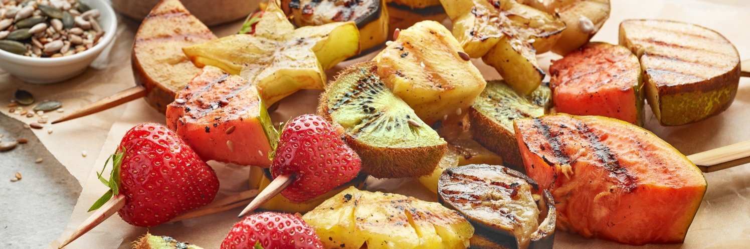 : Gegrillte Holzspieße mit Erdbeeren und Stücken von Kiwi, Birne, Melone, Ananas und Sternfrucht liegen auf Backpapier auf einem Tisch, daneben eine Schale mit Kürbis- und Sonnenblumenkernen.