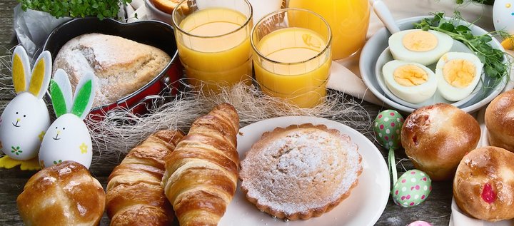 Das Bild zeigt einen Tisch mit Croissants, Muffins, halbierten gekochten Eiern, einem herzförmigen Napfkuchen, einem Törtchen und Gläsern voller Orangensaft, sowie grünen gemusterten Deko-Eiern und Deko-Gras.