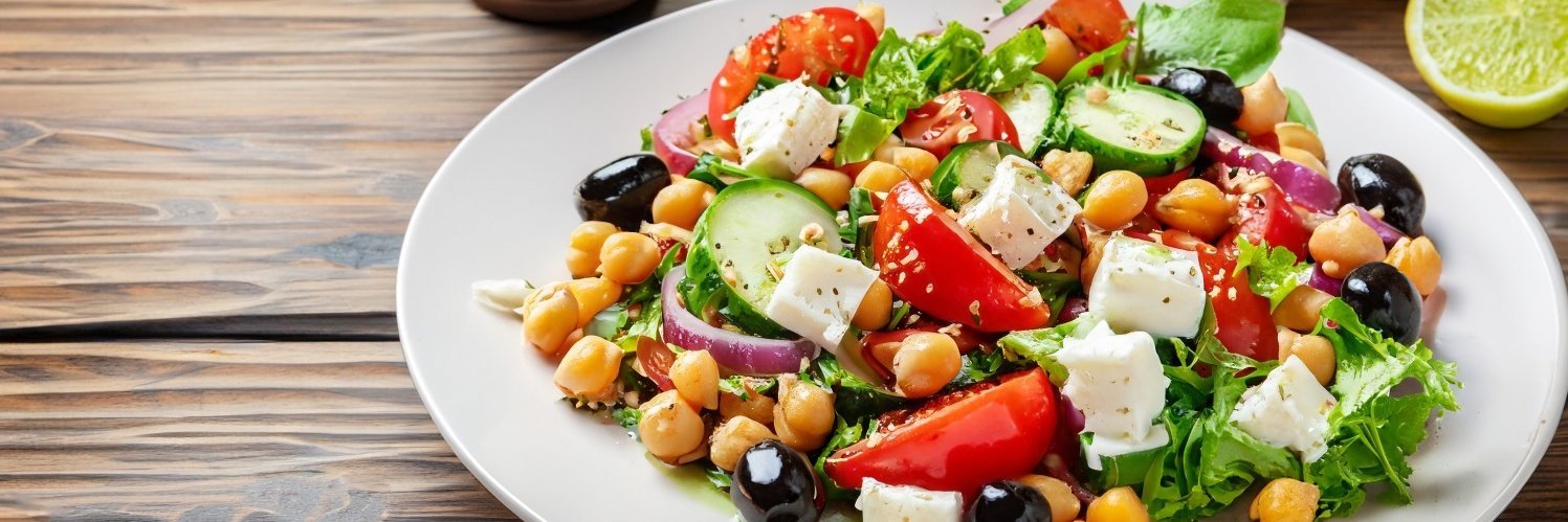 Auf einem dunklen Holztisch steht ein weißer Teller mit Blattsalat, Tomaten, Fetawürfeln, schwarzen Oliven und Kichererbsen.