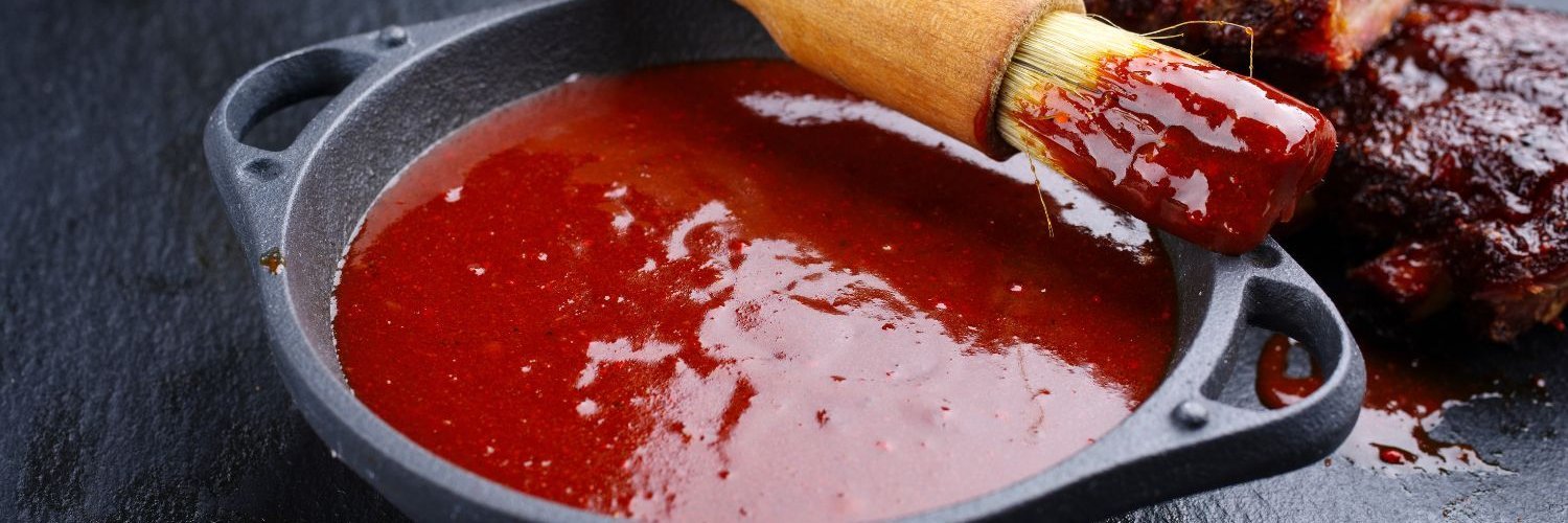 In einer dunklen Schale auf einem Schiefertisch ist Barbecue-Soße und ein Pinsel. Daneben liegen marinierte Spareribs.