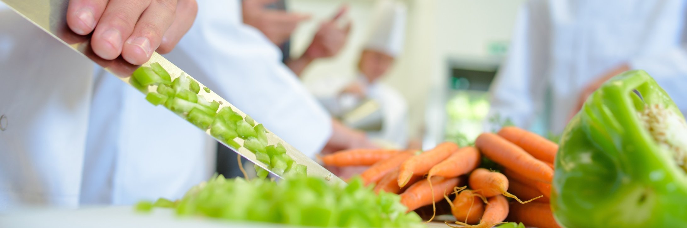 Köche beim Schneiden von Gemüse, im Vordergrund Möhren und Paprika und eine Hand mit scharfem Messer 
