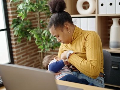 Eine Frau sitzt vor ihrem Laptop und stillt ihr Baby. Sie schaut ihr Baby an.