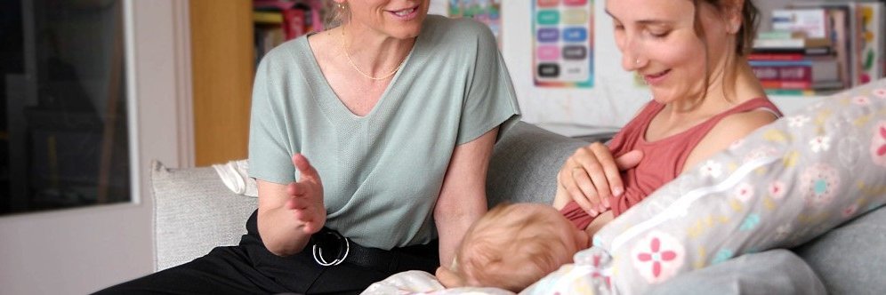 Zwei Frauen sitzen auf dem Sofa, eine stillt ein Baby. Die andere spricht mit ihr.