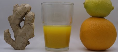 Ein großes Stück Ingwer, ein Zitronen-Orangen-Männchen und ein Glas mir einem kräftig gelben Shot darin