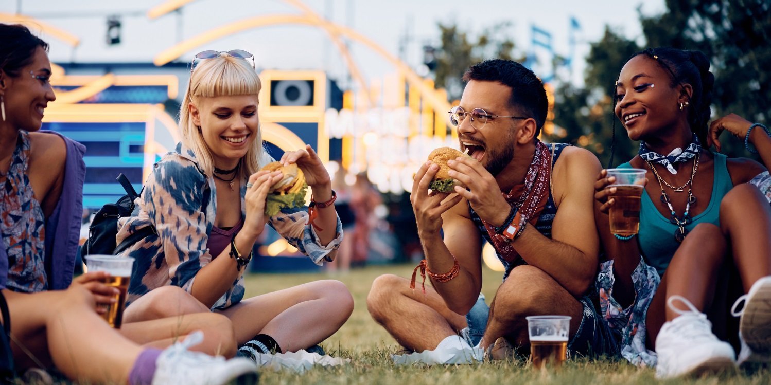 Eine Gruppe junger Erwachsener sitzt auf einer Wiese mit einer Bühne im Hintergrund und isst belegte Brötchen.. Artikel "Food fürs Festival" öffnen.