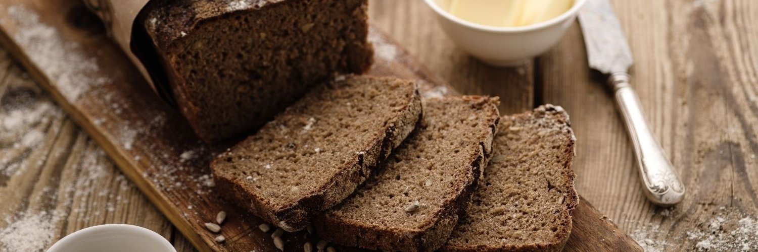 Auf einem Holzbrett liegt ein eckiges dunkles Roggenbrot mit Papiermanschette, von dem 3 Scheiben abgeschnitten sind. Daneben sind Butter, ein Messer und ein Schälchen mit Honig.