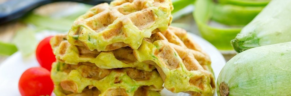 Auf einem Teller liegen mehrere Zucchini-Waffeln. Rechts daneben sind eine Zuchhini und kleine Tomaten. 