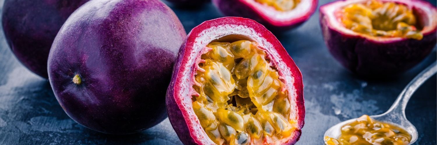Auf einer dunklen Platte liegen ganze und halbierte violette Passionsfrüchte. Ein Löffel mit Fruchtfleisch liegt daneben. 