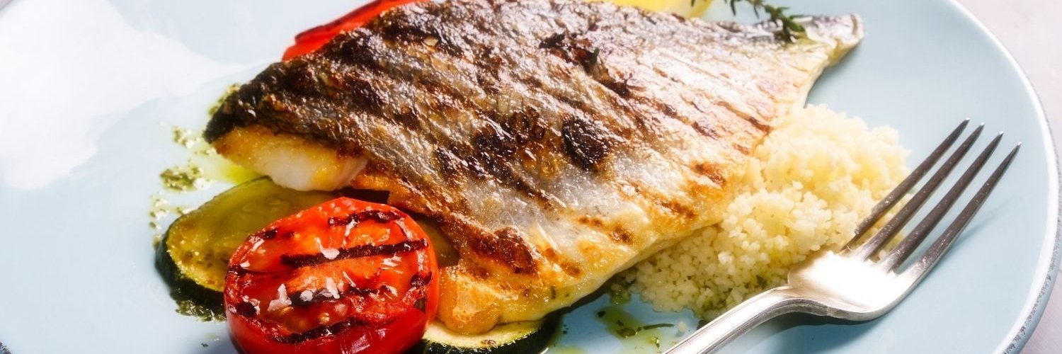 Auf einem weißen Teller liegt ein Stück gegarter Fisch garniert mit Zucchini, Tomate, Kräutern und Couscous neben einer Gabel.
