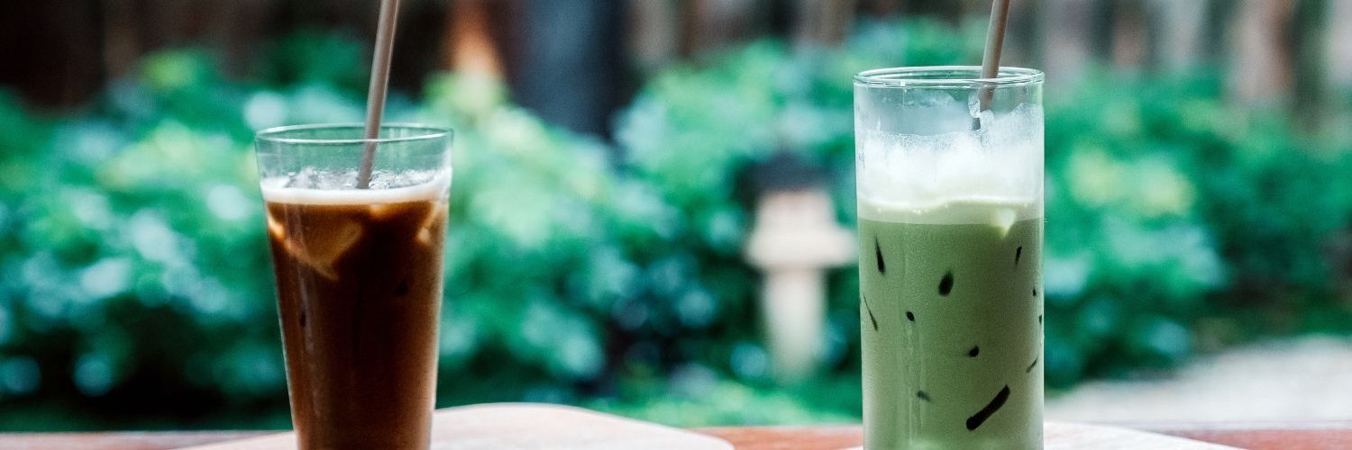 Ein Glas mit Hojicha-Eistee und ein Glas Iced Matcha-Latte stehen nebeneinander auf einem Holztisch.