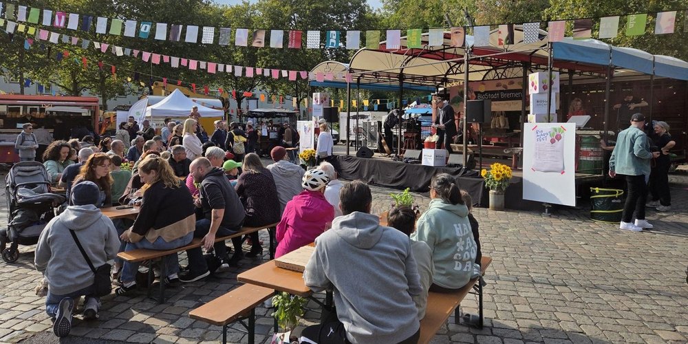 Besucherinnen und Besucher des BioMarktFestes sitzen an Biertischen vor einer Bühne.