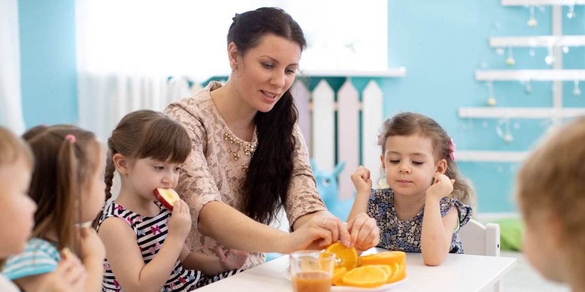 Kitakinder essen mit ihrer Erzieherin vorgeschnittene Orangen. . Artikel "Professionelle Haltung beim Essen" öffnen.