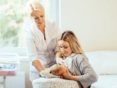 Eine junge Frau legt ihr Baby an die Brust. Eine Hebamme hilft beim Positionieren des Babys.