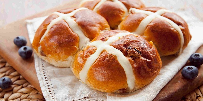 Auf einem Holzbrett liegen vier hellbraune weich aussehende Brötchen. Jedes hat ein helles Kreuz aus hellem Teig eingebacken. Als Dekoration ein Deckchen und Blaubeeren.. Artikel "Osterspezialitäten aus aller Welt" öffnen.